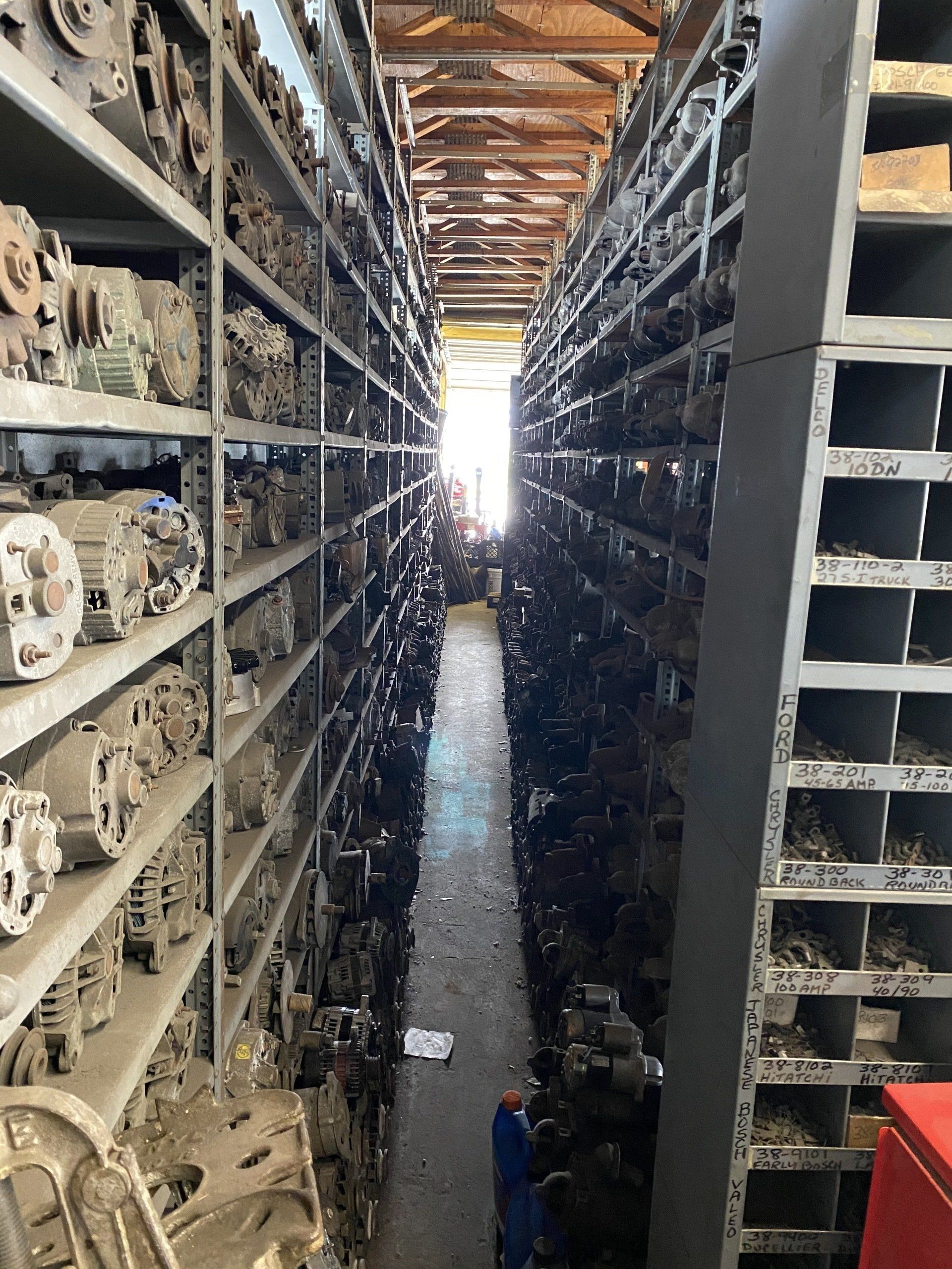 Narrow aisle between tall shelves packed with mechanical parts in a warehouse, bright light at the far end