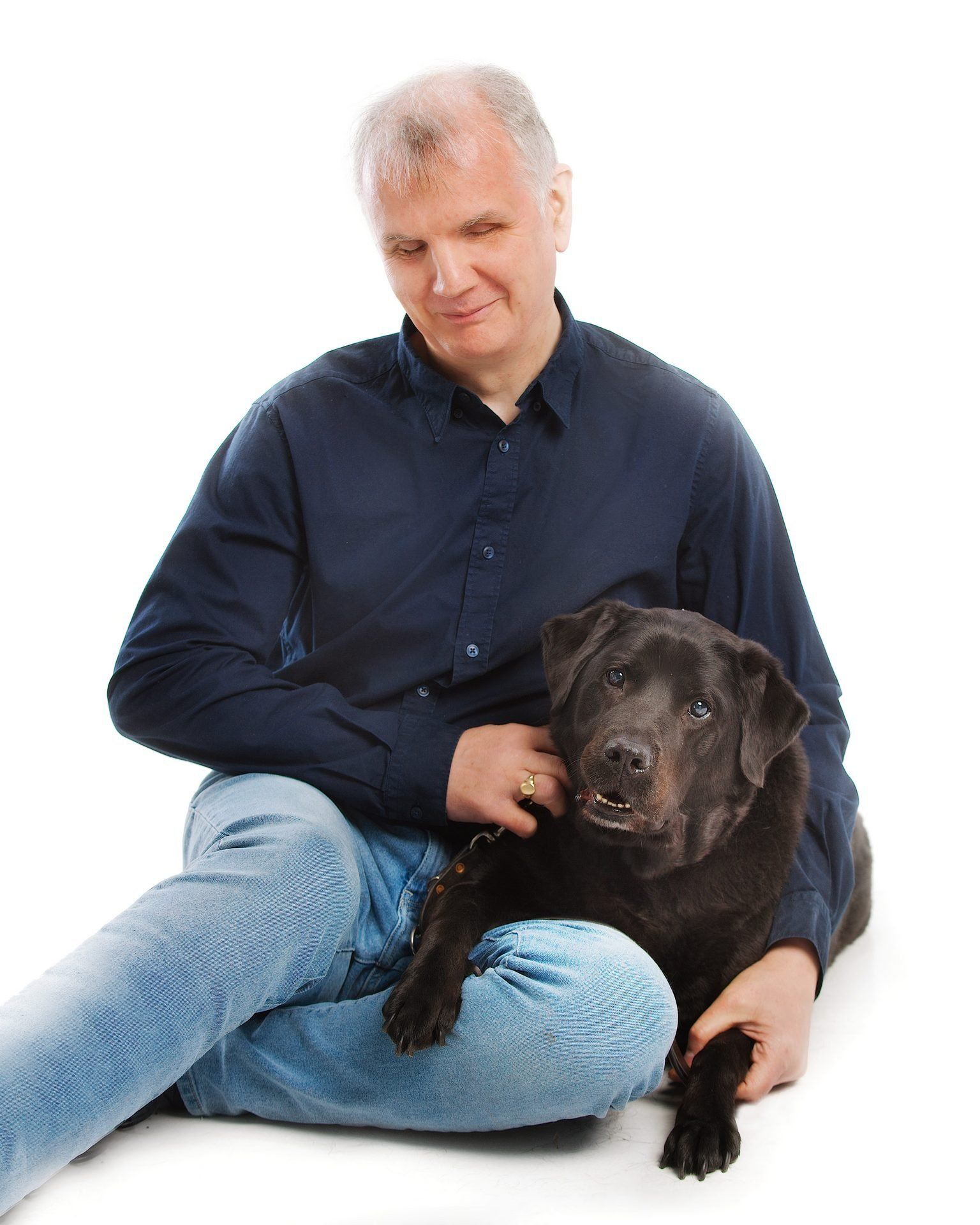 Informal pose of Philip Anderson sitting on the floor with one arm around his former black guide dog, Quintana, who has one paw resting on his knee