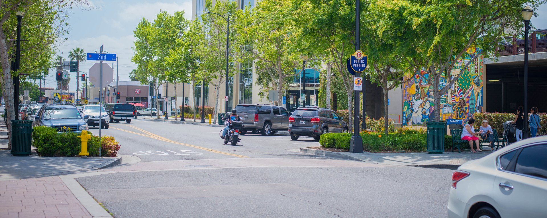 City street scene with cars, trees, and a motorcycle. People are seated on a bench on the right.
