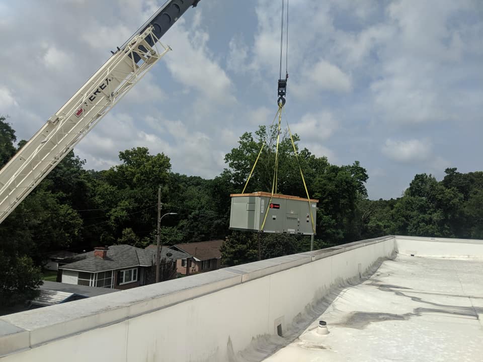 Crane lifting an HVAC unit onto a flat white roof, with trees and houses in the background.