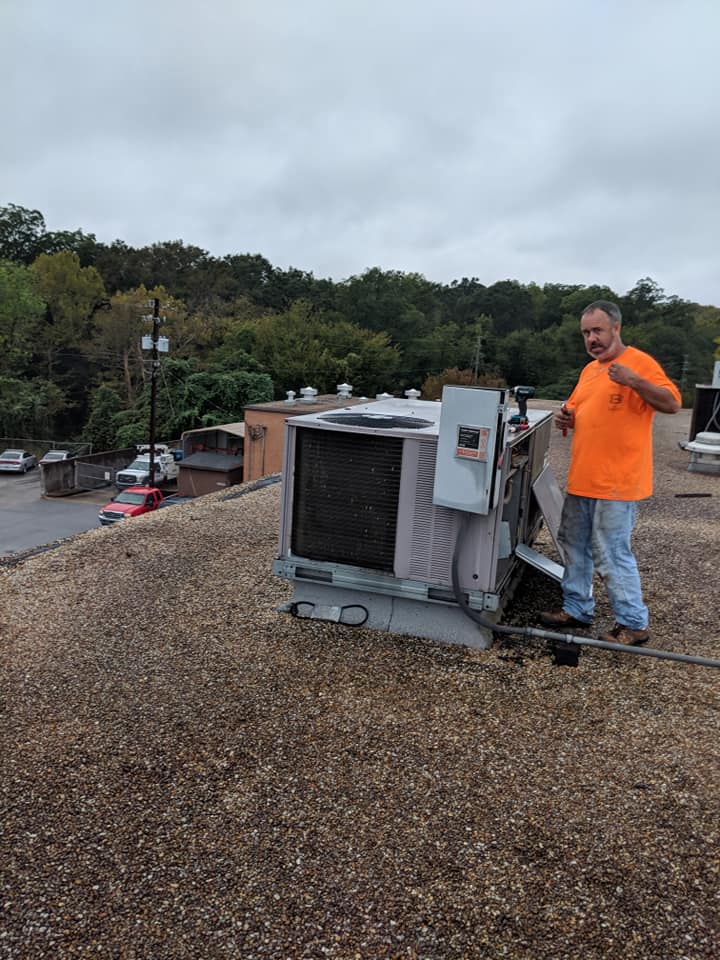 Man in orange shirt on rooftop, near HVAC unit, pointing. Gray skies, gravel roof.