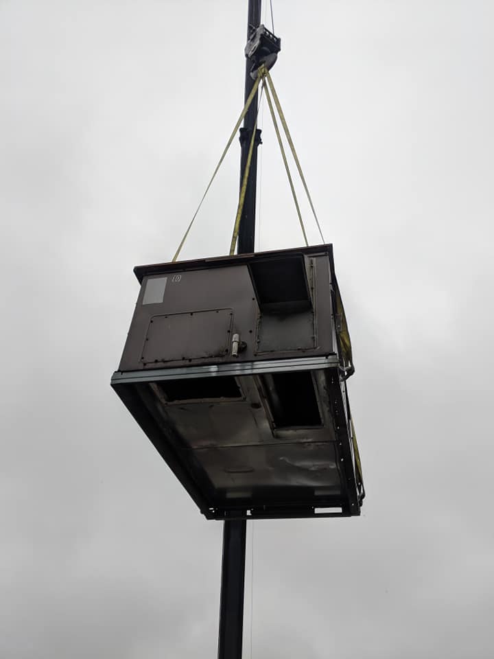 Crane lifting a large crate, two workers guiding it. Cars parked nearby in an asphalt lot.
