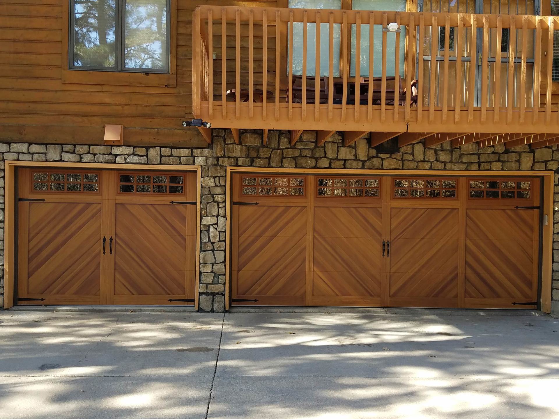 Three wooden garage doors with a chevron pattern and upper windows, set in a stone and wood house with a balcony above.
