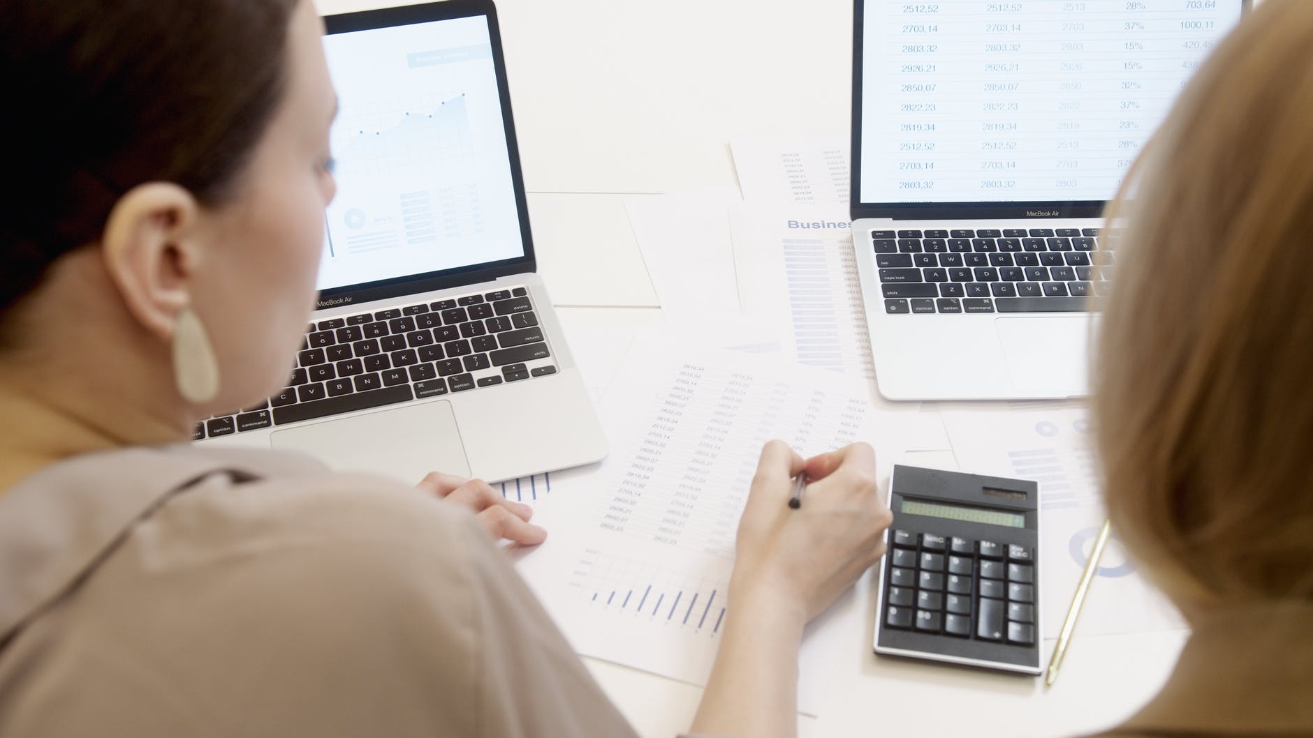Two people working on laptops and paperwork with a calculator.