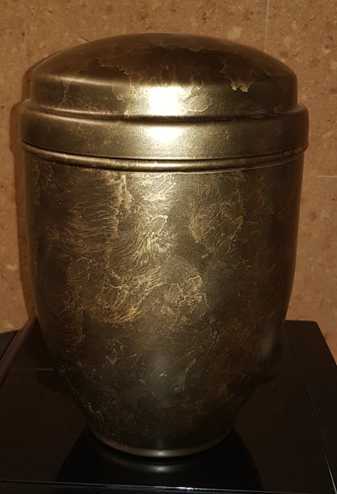 Urna dorata con coperchio arrotondato, appoggiata su una superficie nera su sfondo marrone.