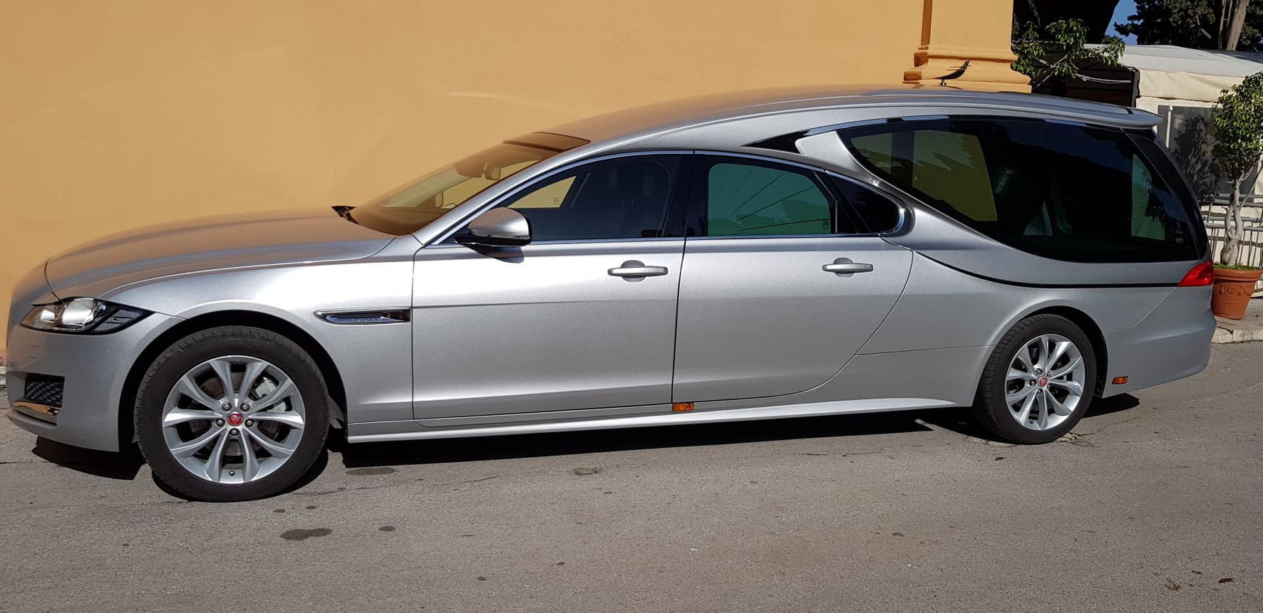 Carro funebre Jaguar argentato parcheggiato accanto a un muro giallo.