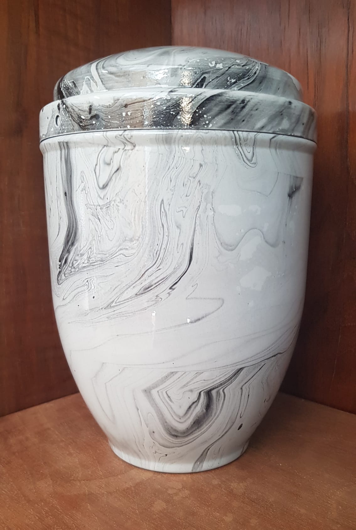 Urna in ceramica bianca e grigia con motivo marmorizzato e coperchio, posta su una mensola in legno.