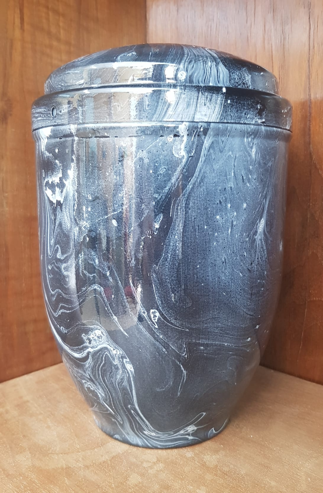 Urna cineraria marmorizzata grigia e bianca con coperchio, appoggiata su una superficie di legno.