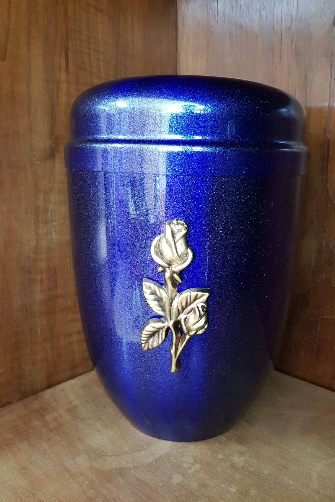 Urna blu con finitura glitterata e decorazione a forma di rosa dorata, posta su una mensola in legno.