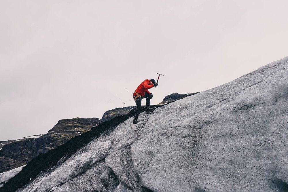 Person in red jacket ice climbing with an ice axe on a glacier, overcast sky.