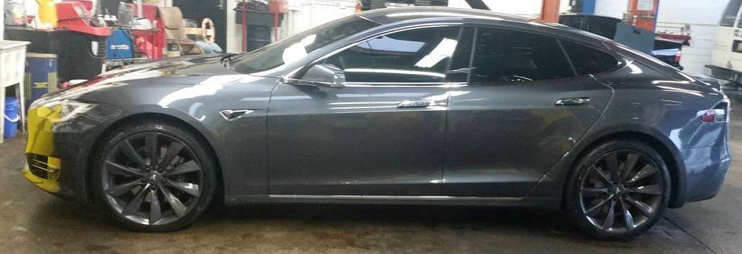 Gray Tesla sedan with tinted windows parked in a garage. Yellow tape covers the front bumper.