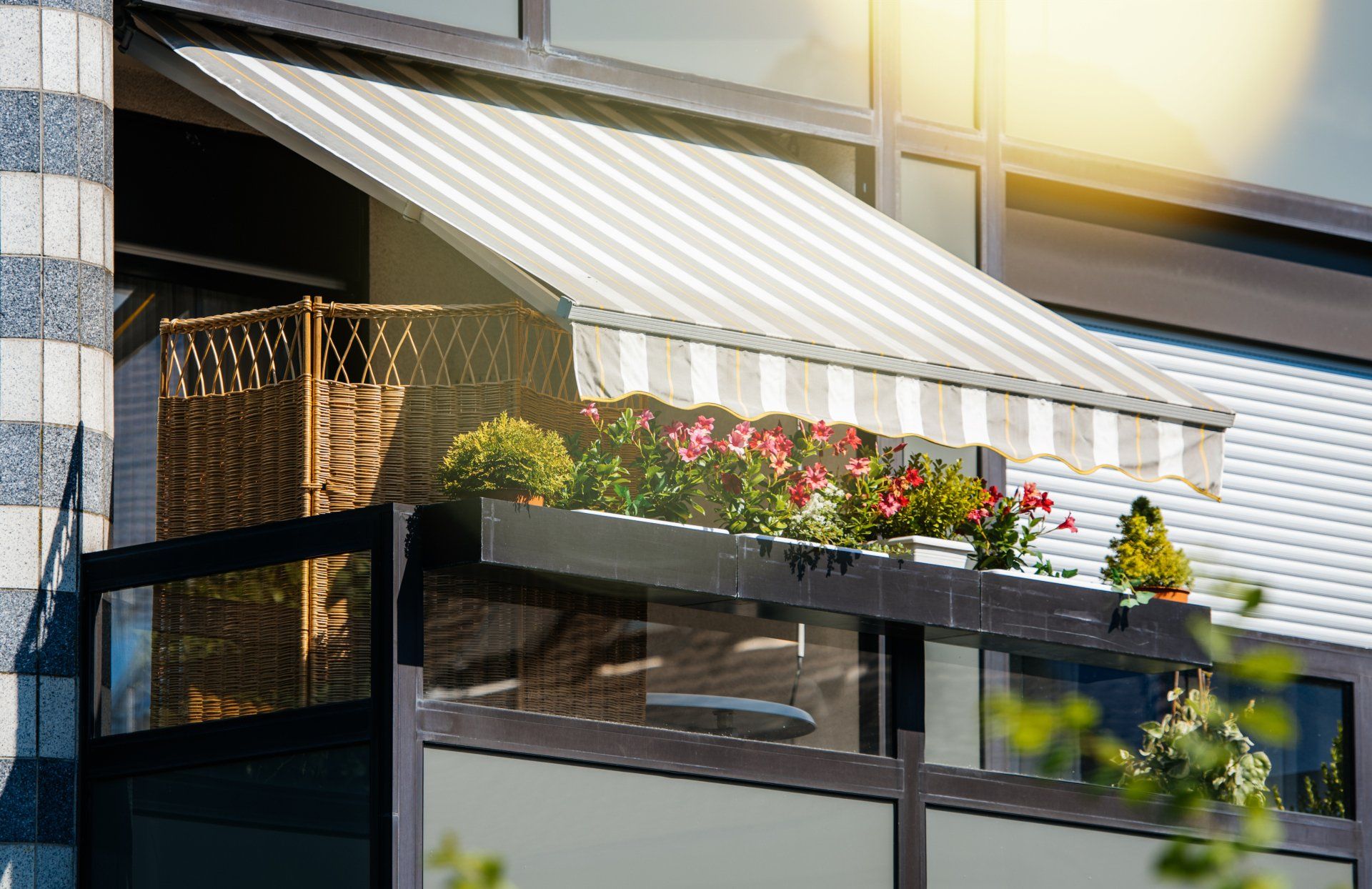 Striped outdoor awning extending over balcony with cane divider, and pink flowers in pots along railing.