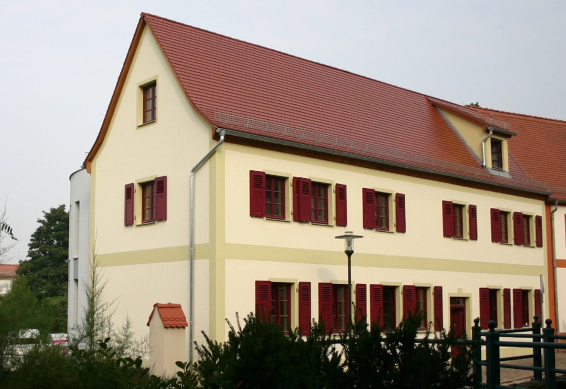 Cremefarbenes Haus mit rotem Dach und Fensterläden, vom Vorgarten aus gesehen