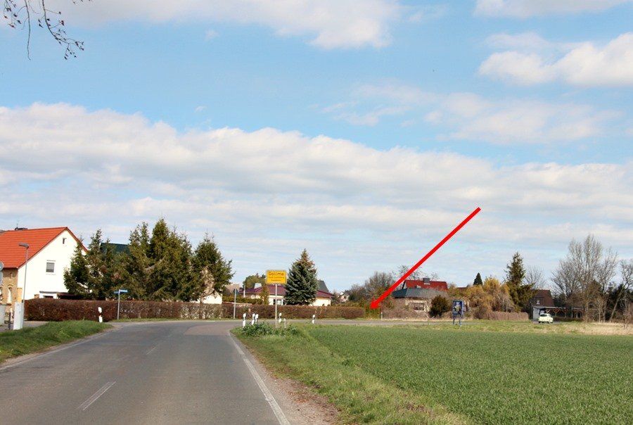 Ländliche Straße neben grünen Feldern und Häusern, ein roter Pfeil weist auf ein entferntes Gebäude.