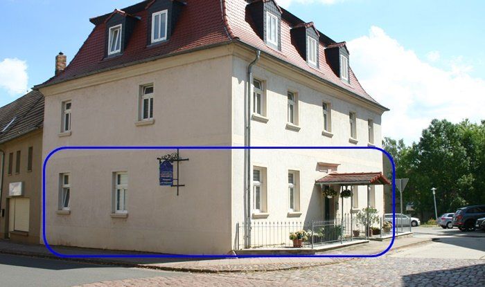 Die blau umrandete Seite eines cremefarbenen Hauses mit Fenstern und einer kleinen Terrasse an einem Kiesplatz.