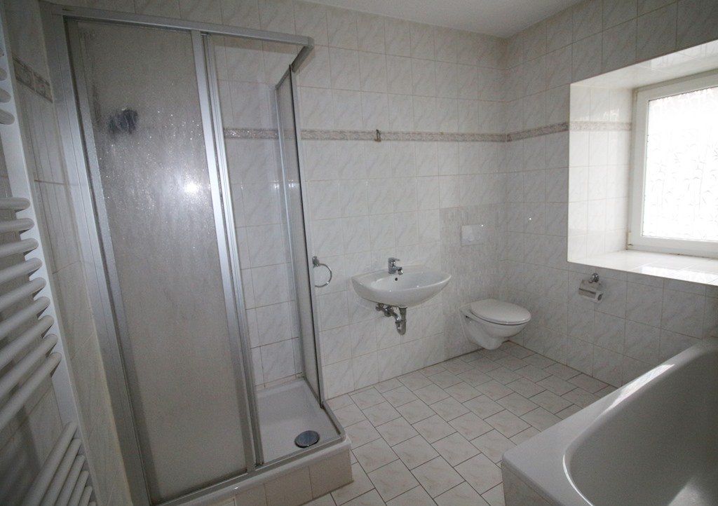 Kleines weißes Badezimmer mit Dusche, Waschbecken, Toilette und gefliestem Boden in der Nähe eines hellen Fensters.