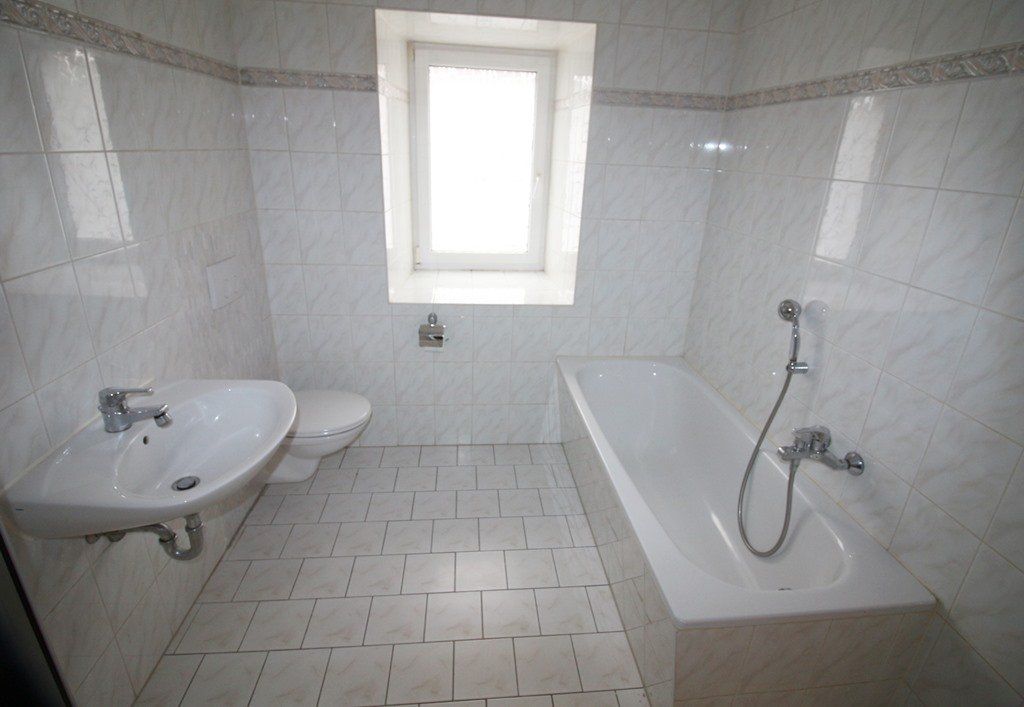 Kleines, weiß gefliestes Badezimmer mit Waschbecken, Toilette, Badewanne und Fenster.