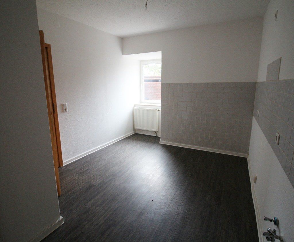 Leerer weißer Raum mit dunklem Holzboden, grauen Wandpaneelen und einem Fenster mit einem Heizkörper darunter.