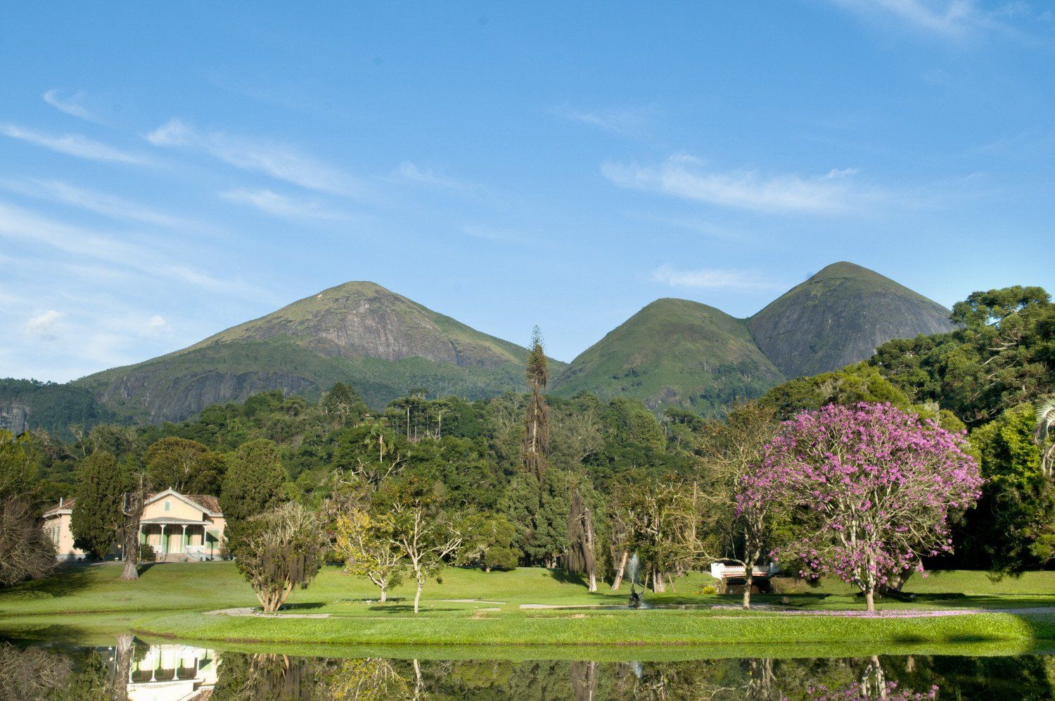 Paisagem verdejante com montanhas e lago sob um céu azul. Edifícios e árvores em primeiro plano, reflexos na água.