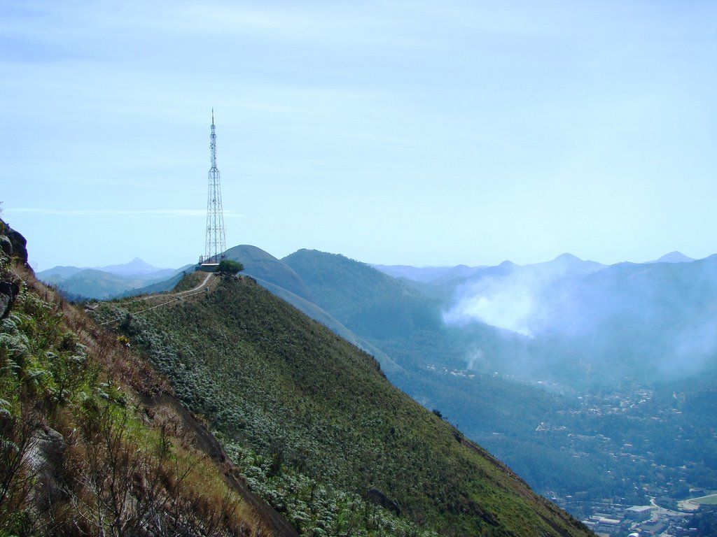 Crista de montanha com torre de comunicação, com vista para um vale envolto em névoa ao longe e uma cidade sob um céu azul.