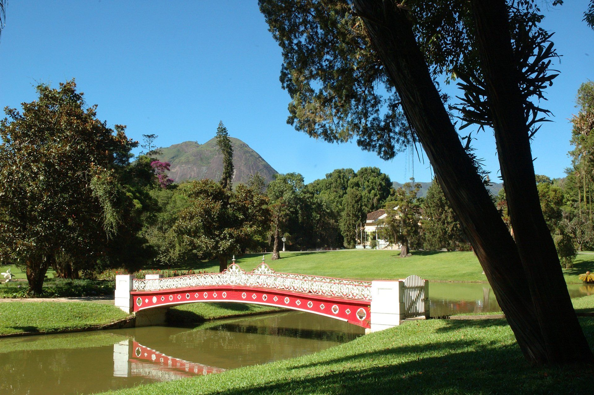 Ponte vermelha e branca sobre a água em um parque, com árvores e uma montanha ao fundo.