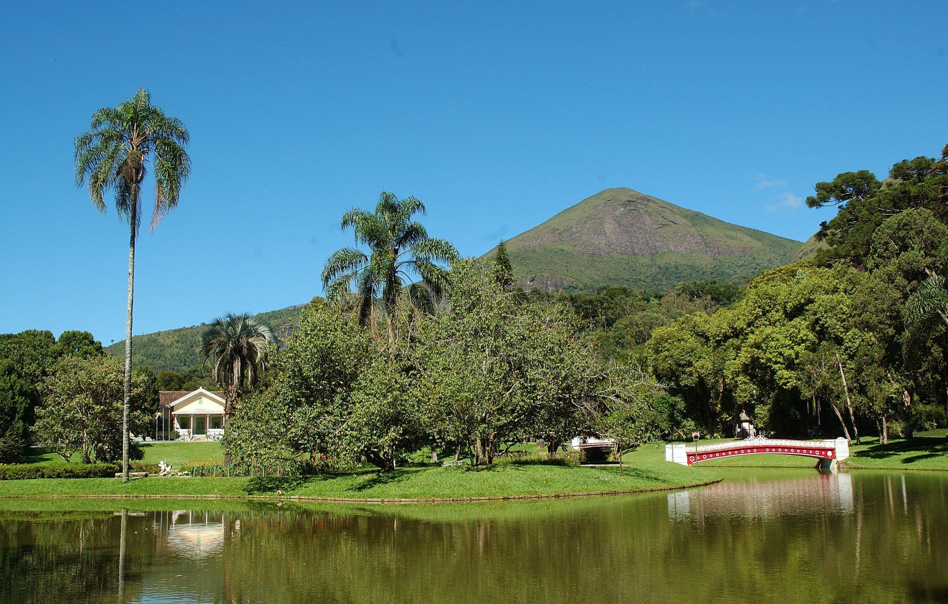 Paisagem verdejante com lago, ponte, casa e montanha sob um céu azul límpido.