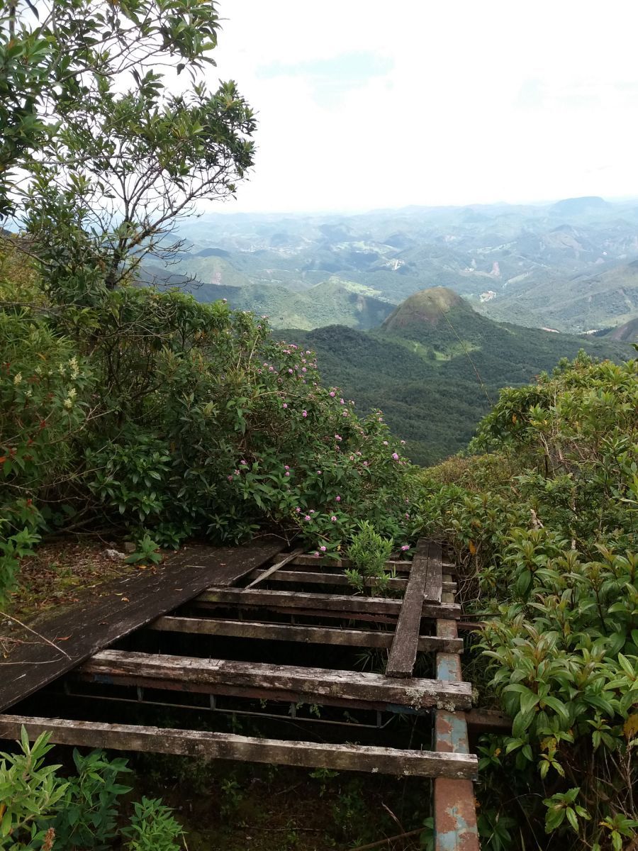 Degraus de madeira descem por uma trilha na montanha, com um vale verdejante ao longe visível.