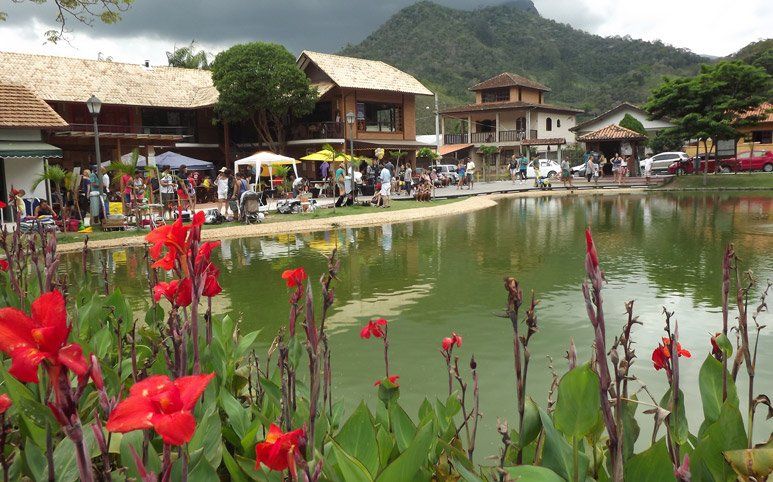 Lago exuberante com flores vermelhas em primeiro plano, lojas e pessoas ao redor, montanhas ao fundo.
