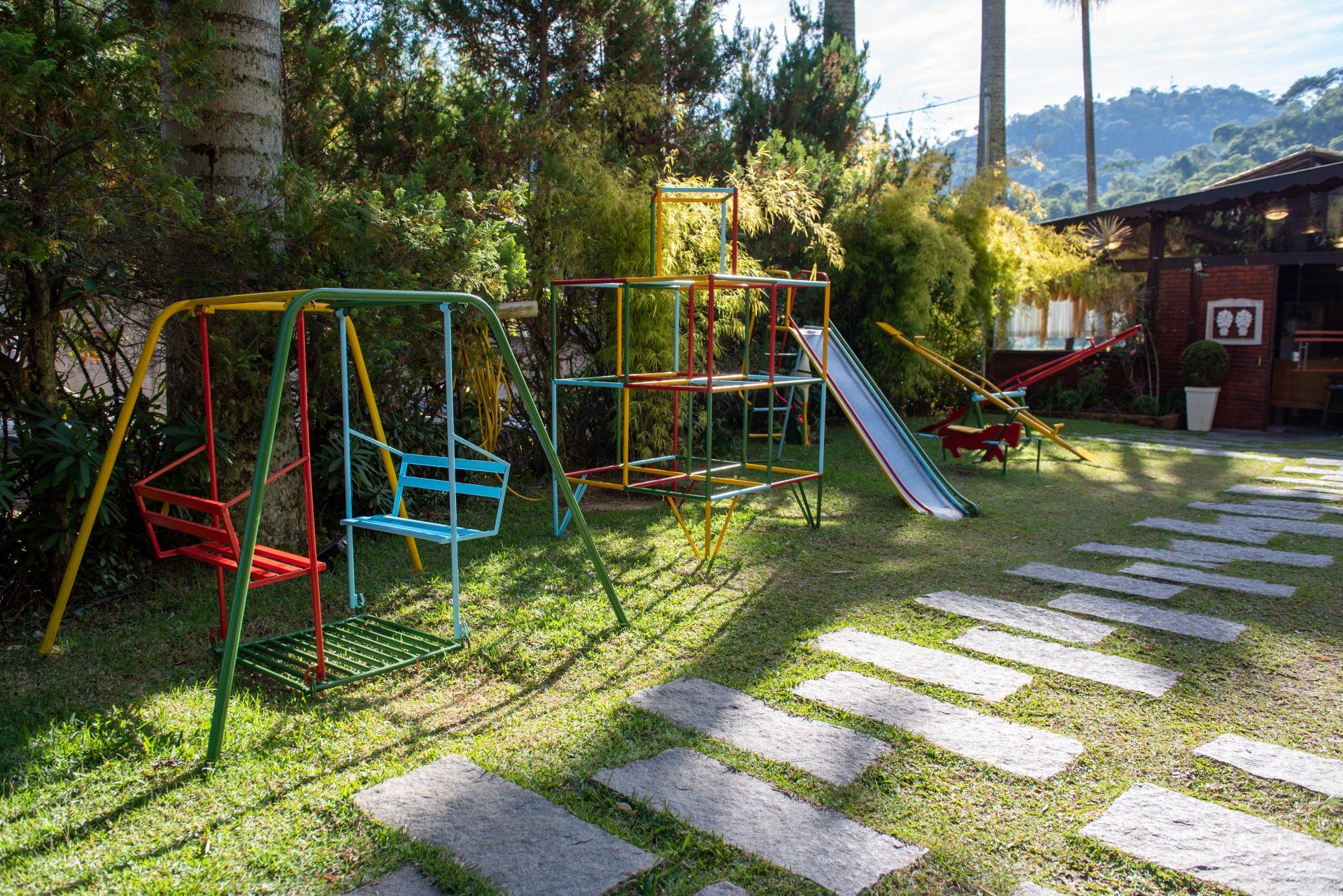 Parque infantil com balanços, estrutura de escalada e escorregador em um gramado com caminho de pedra.
