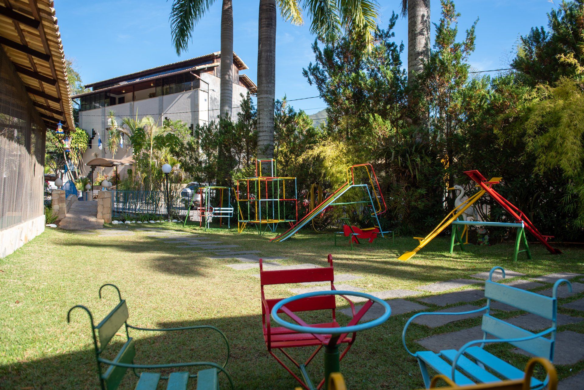 Parque infantil com escorregadores, balanços e cadeiras coloridas em um gramado. Prédio ao fundo.