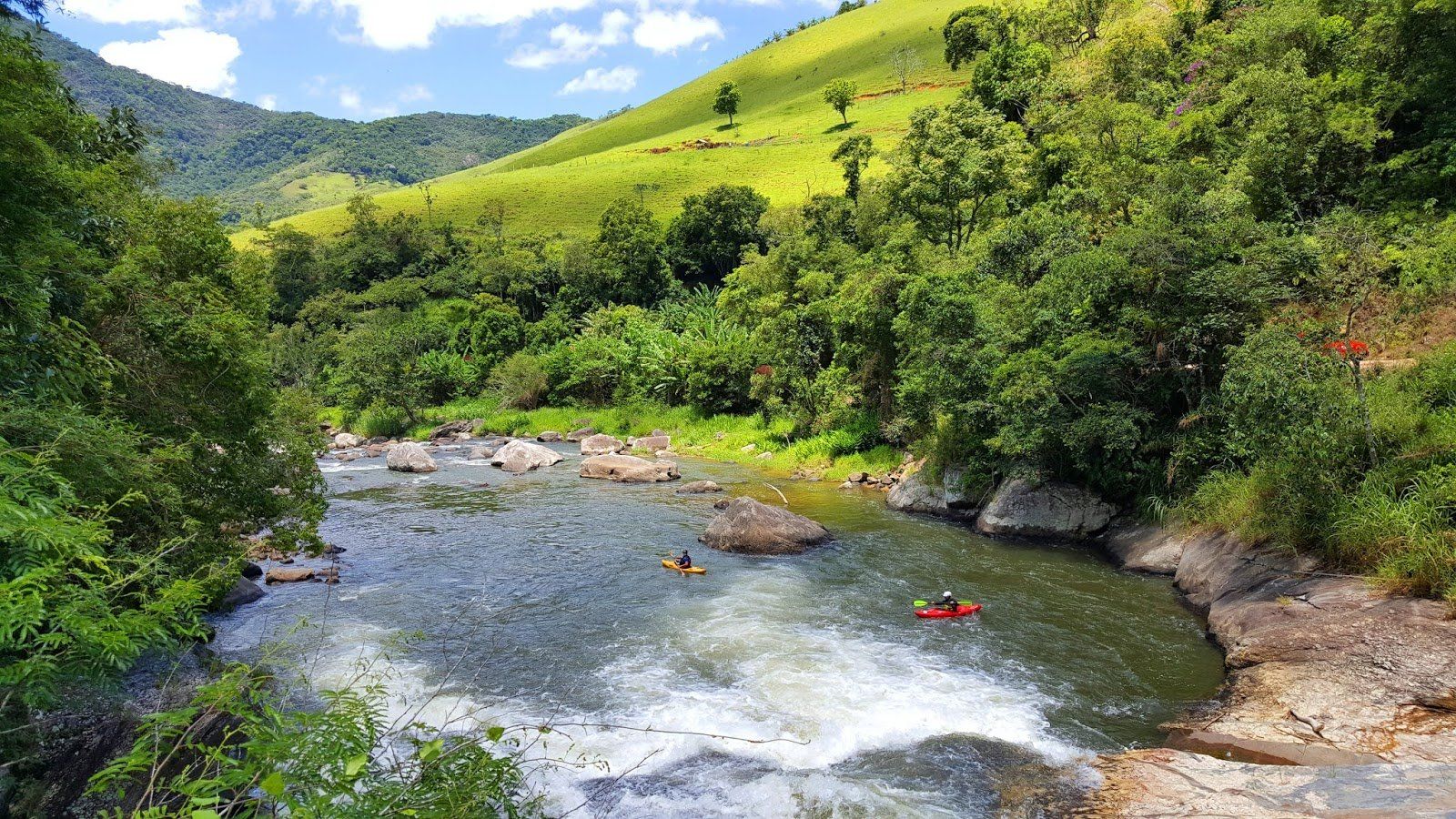 Rio serpenteando por colinas verdejantes, caiaques na água, árvores exuberantes margeando as margens.