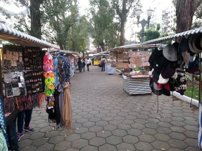 Mercado ao ar livre com barracas vendendo joias, lenços e chapéus em um caminho pavimentado, com pessoas passando.