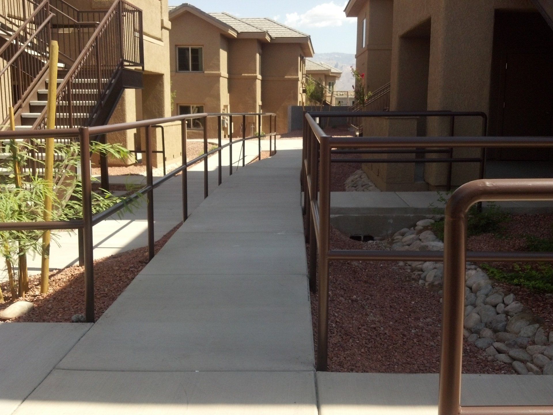 Concrete ramp with brown metal handrails in front of a tan apartment building, red gravel landscaping.
