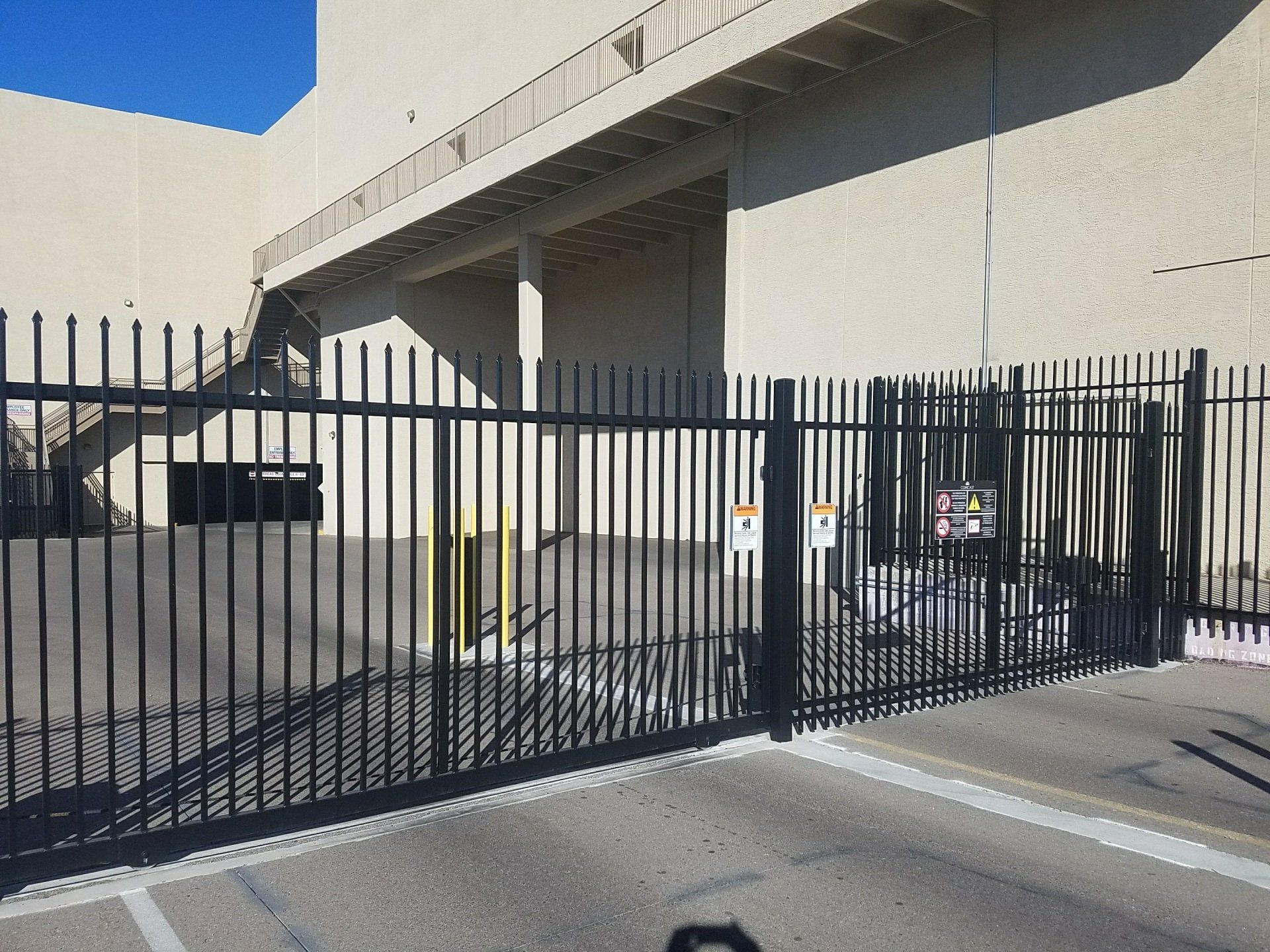 Black metal gate in front of a tan building. Concrete parking area. Shadows cast.