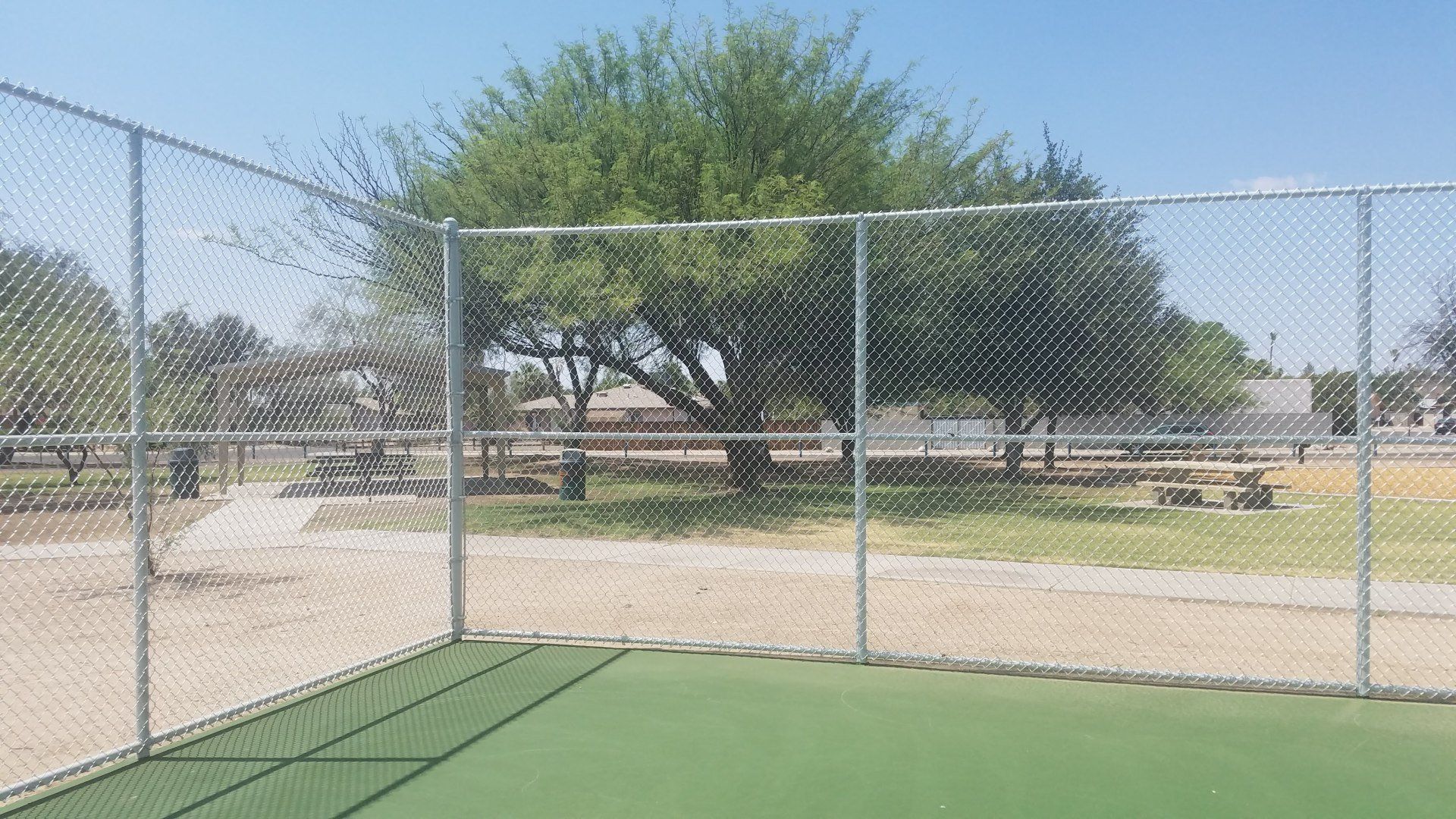 Chain-link fence surrounding a green surface, looking into a park with picnic tables and a tree.