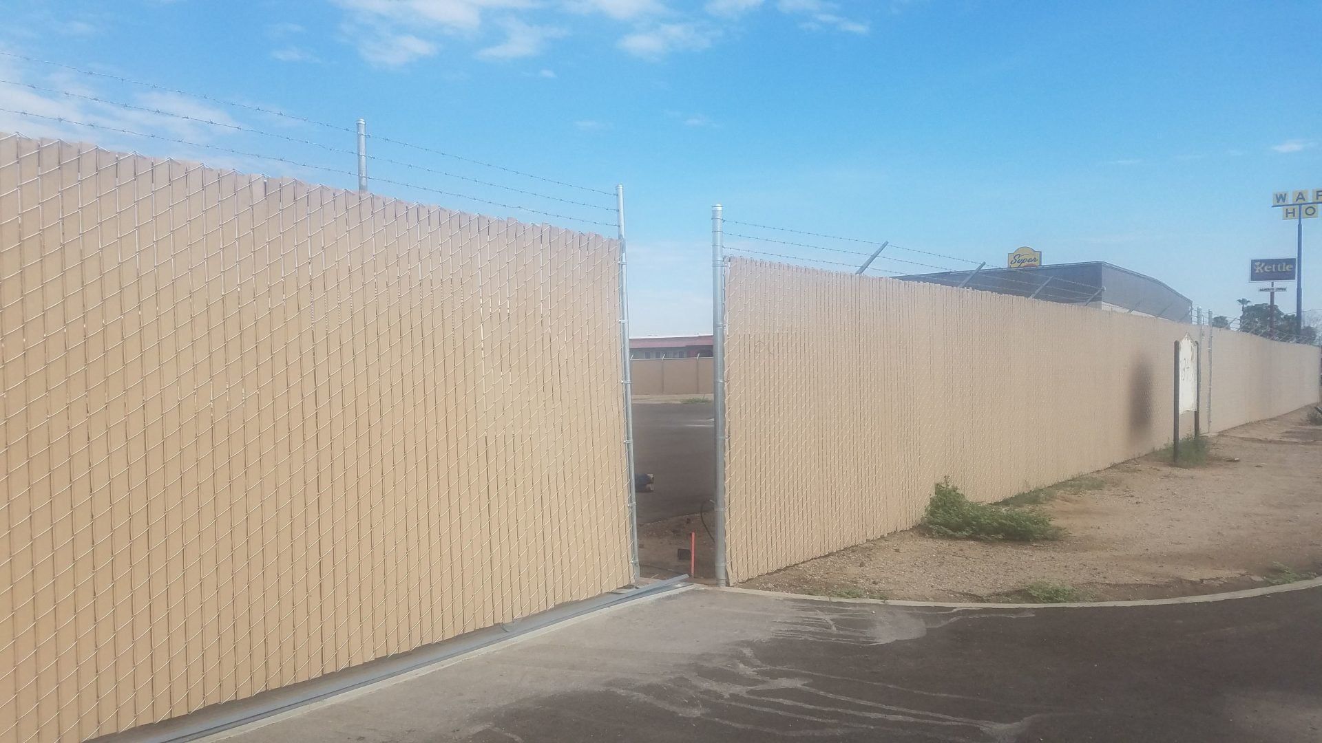 Tan slatted security fence with razor wire, open entrance, blue sky.