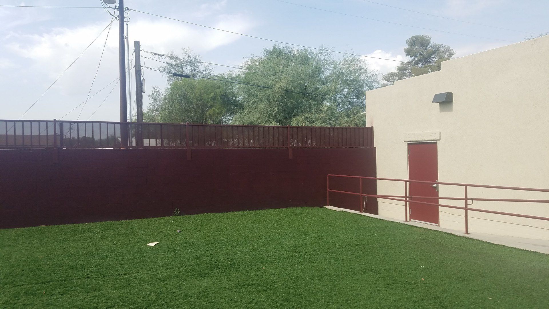 Green lawn, maroon fence, tan building with a red door, and trees against a cloudy sky.