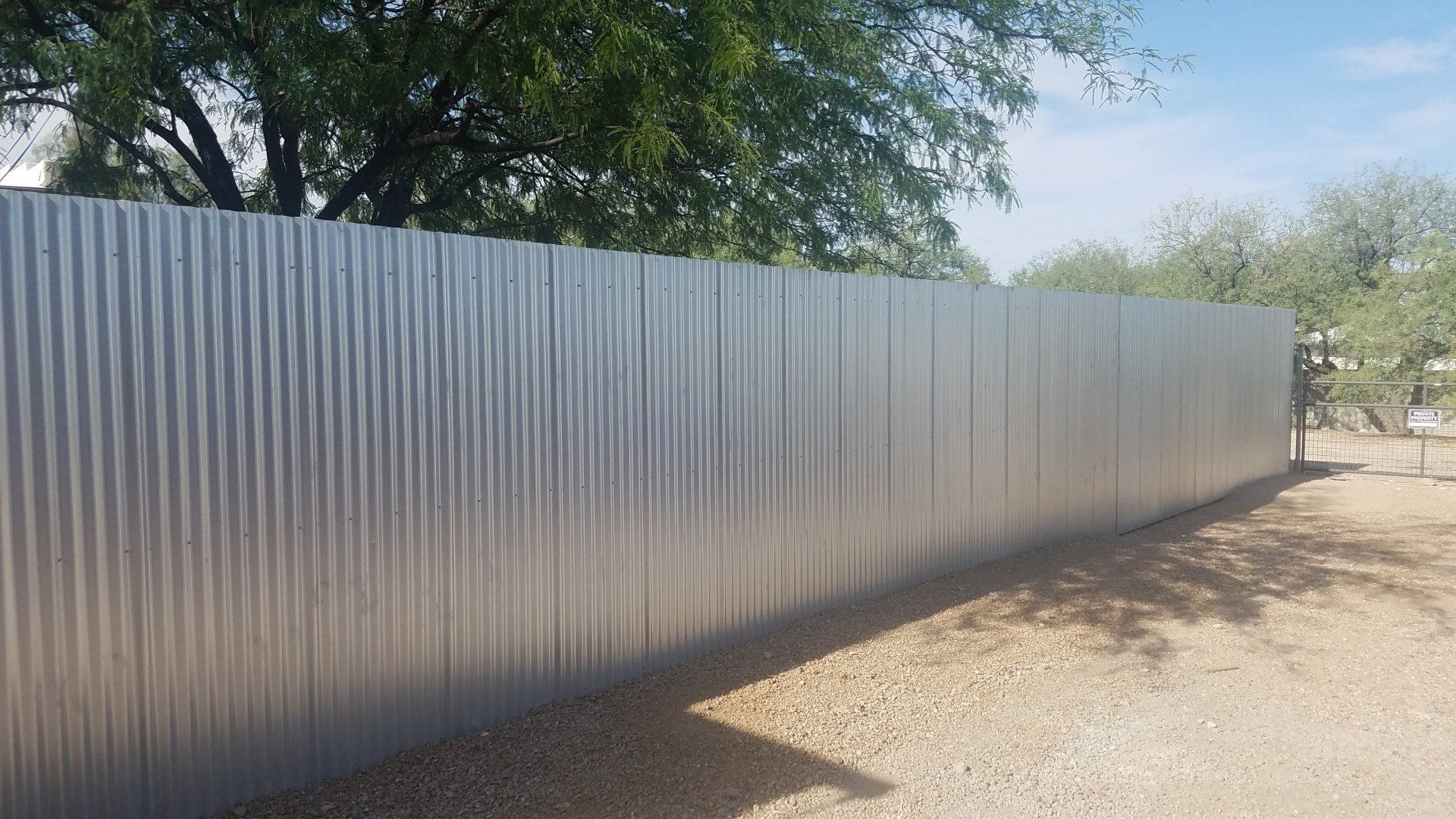 Corrugated metal fence on gravel in front of trees, under a blue sky.