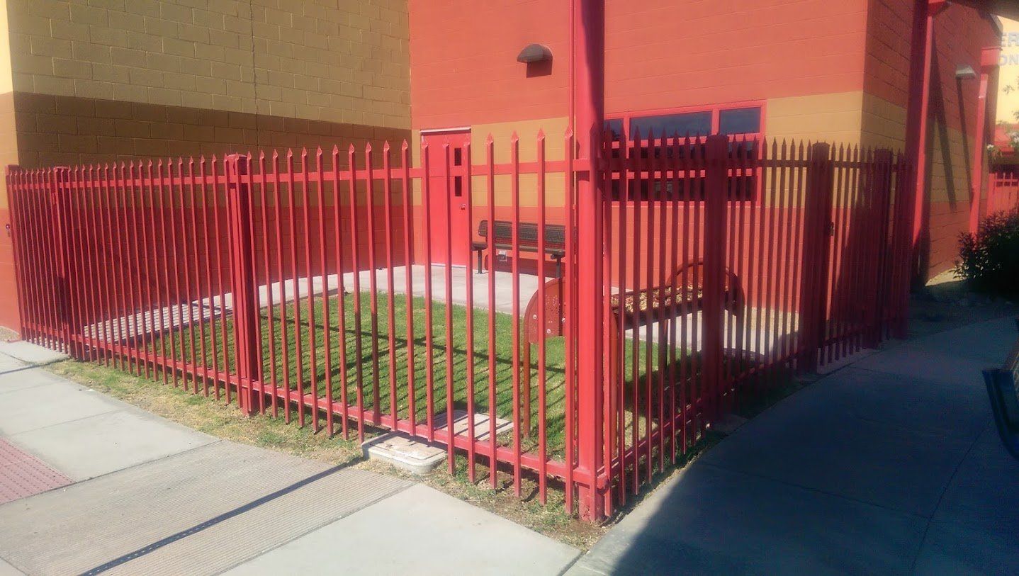 Red metal fence encloses a small, grassy area next to a building. Sidewalk in the foreground.