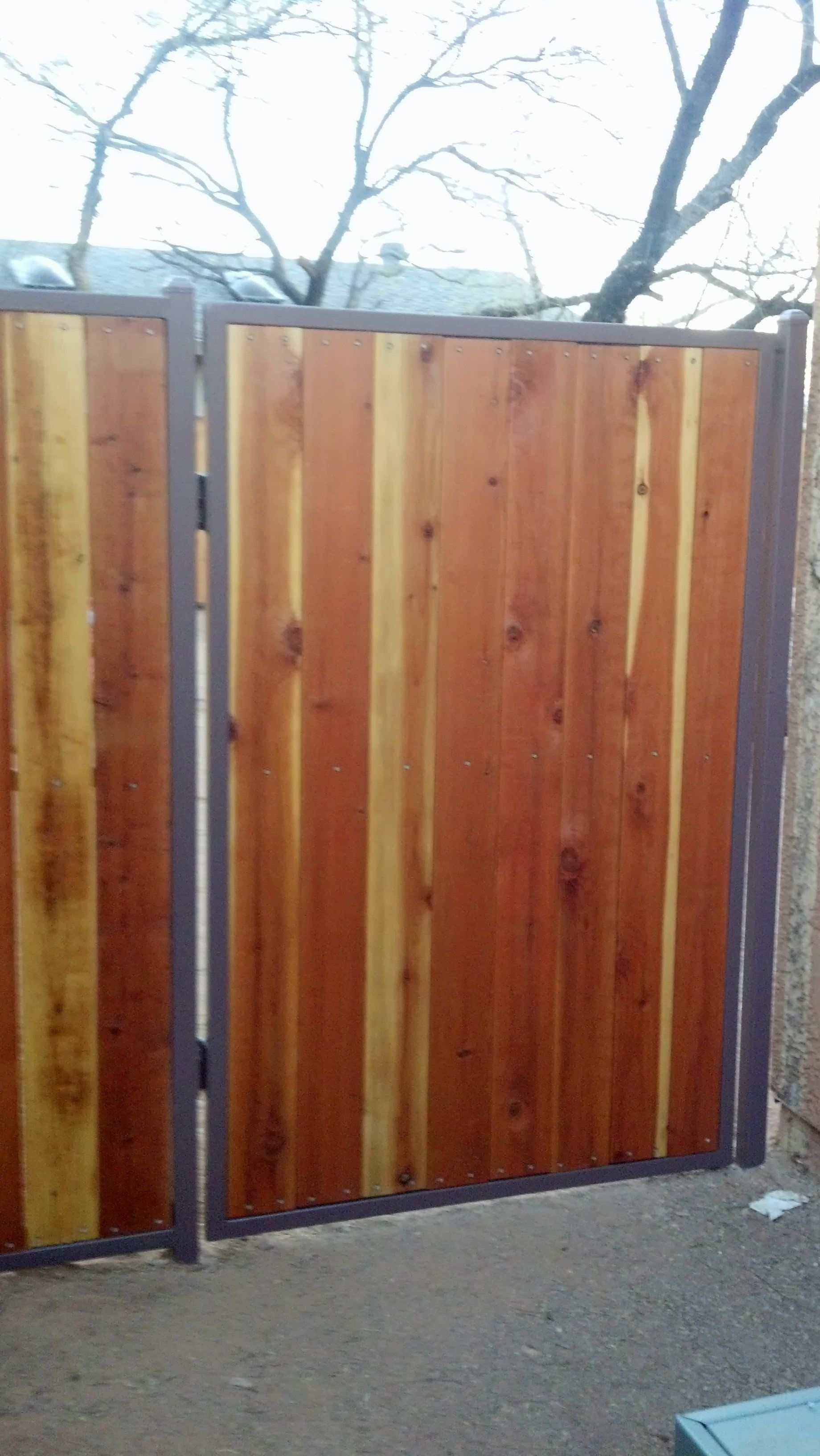 Wooden gate with vertical planks, framed in dark metal, set against a tree-filled sky.