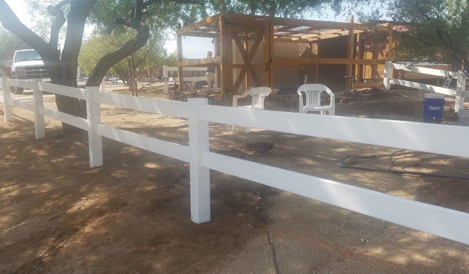 White two-rail fence along a dirt road, with a wooden shed and tree in the background.