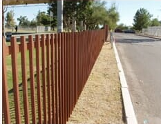 Brown metal vertical slatted fence alongside a road, under a clear sky.