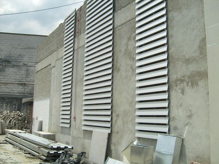 Muro exterior con tres respiraderos altos y verticales con rejillas de ventilación; materiales de construcción en primer plano.