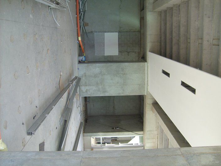 Interior de una obra con escaleras y paredes de hormigón. Un panel blanco con recortes negros recubre el hueco de la escalera.