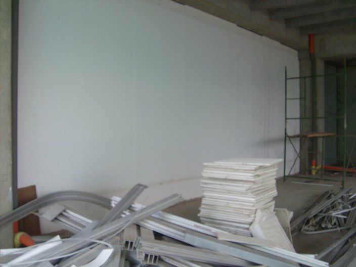 Interior de una obra en construcción con una gran pared blanca, andamios y materiales de construcción en primer plano.