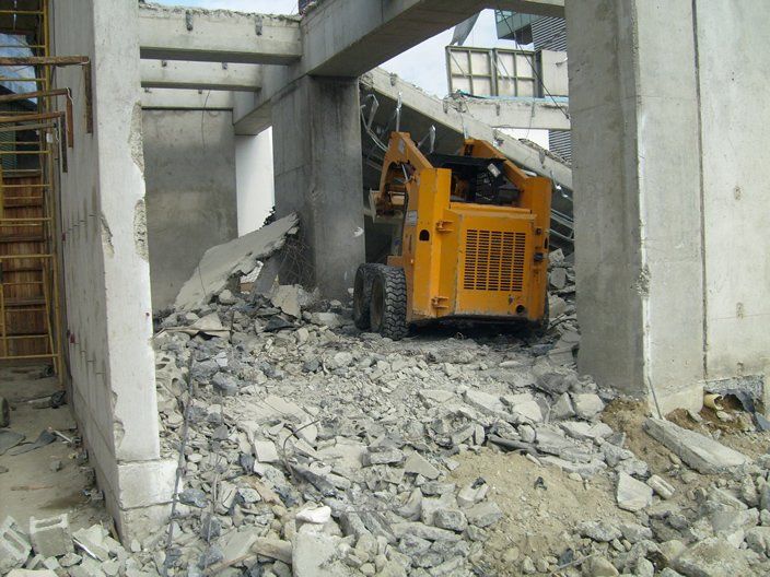 Minicargadora amarilla limpiando escombros de un edificio parcialmente demolido.