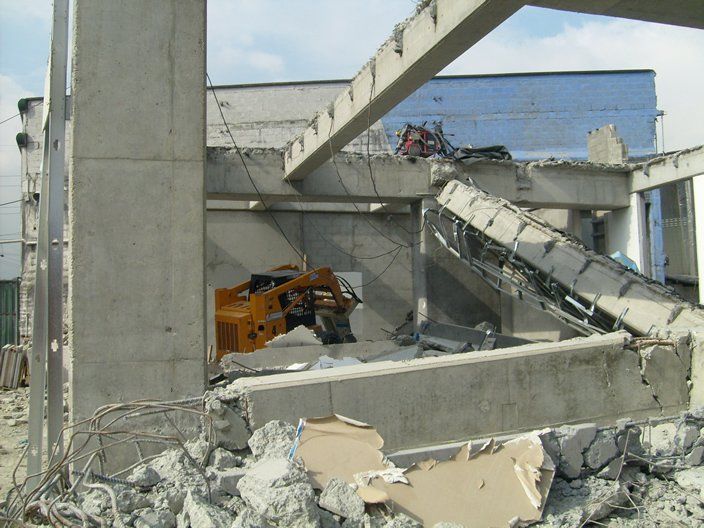 Edificio de hormigón siendo demolido por una excavadora naranja, con escombros esparcidos por todo el lugar.