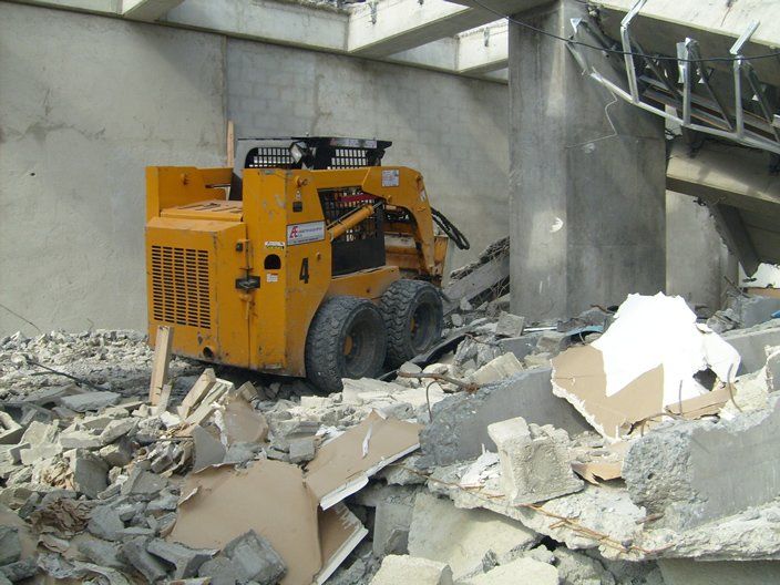 Cargadora compacta amarilla en medio de escombros de demolición dentro de un edificio, posiblemente limpiando escombros.