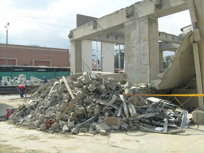 Montón de escombros frente a un edificio parcialmente demolido. Trabajadores al fondo; se ve el cielo.