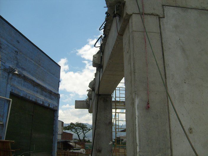 Estructura de hormigón con arco, obra en construcción. Edificio azul a la izquierda, con árboles y cielo al fondo.